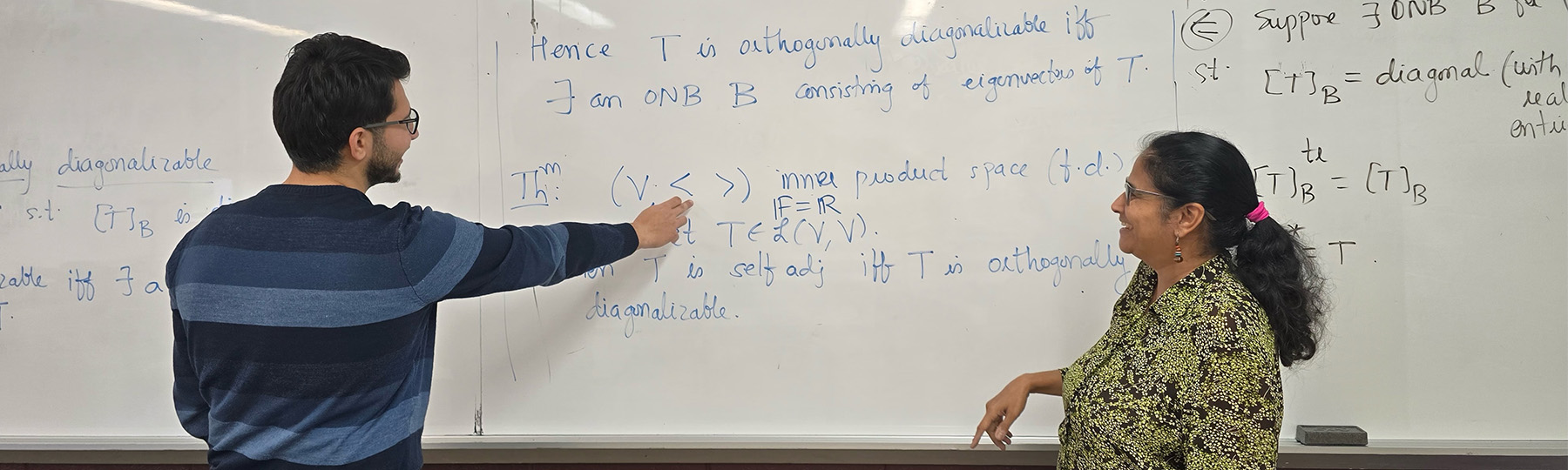 A gradate student wearing a blue striped shirt talks with a graduate professor about the mathematical equation on the white board behind them.