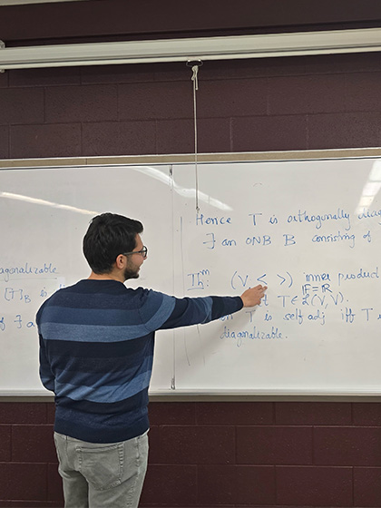 A gradate student wearing a blue striped shirt points out a mathematical equation on the white board behind him.