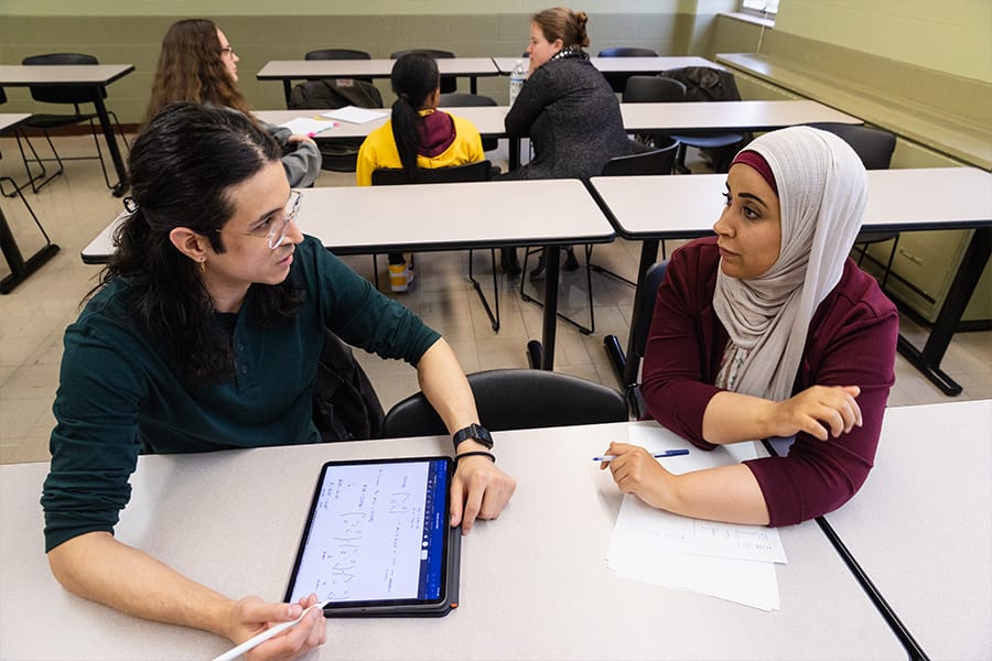 Two pure mathematics students sit at a table discussing an equation displayed on an iPad screen.