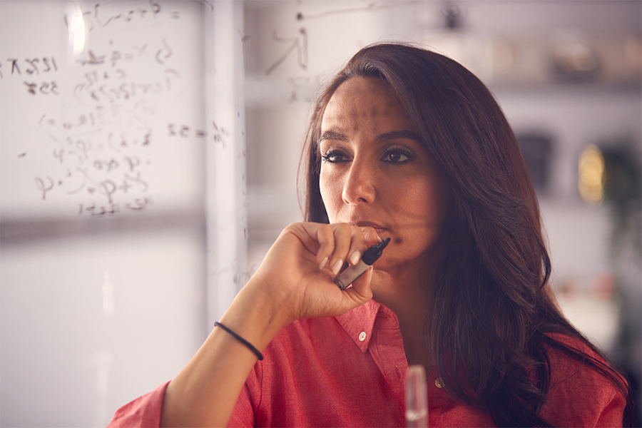 A woman with long brown hair hold a dry erase marker while standing in front of plexiglass solving a mathematical equation.