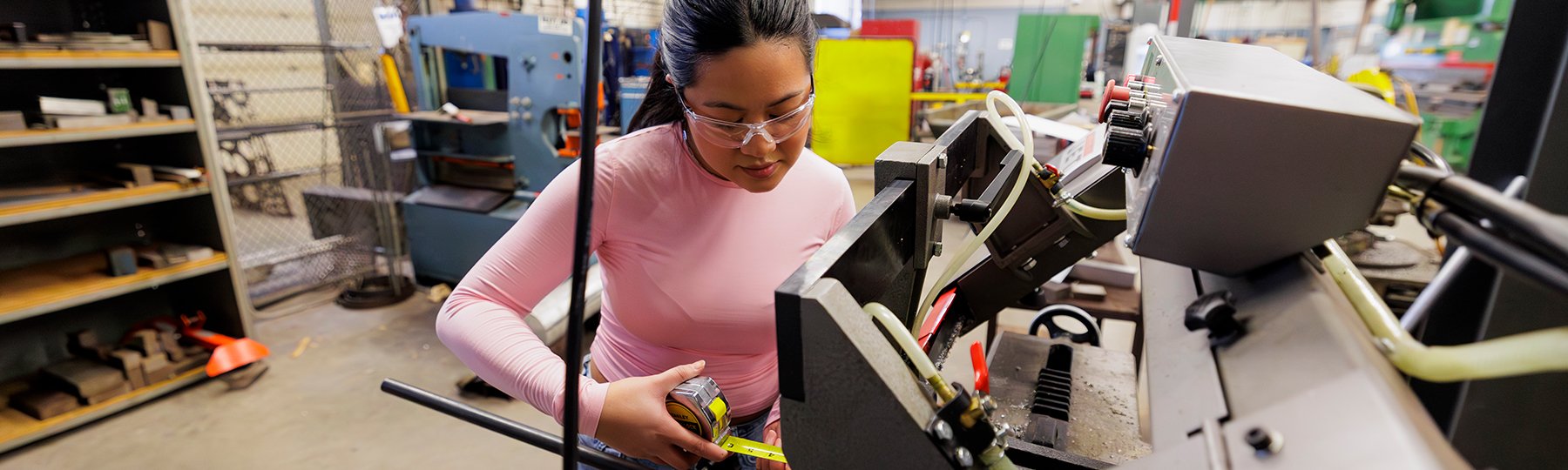 A woman wearing safety goggles using a tape measure while standing in front of machinery equipment.