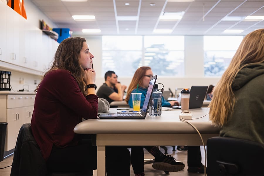 A student in a maroon top sits at a table with a laptop in front of her. There are other students around her in the classroom.