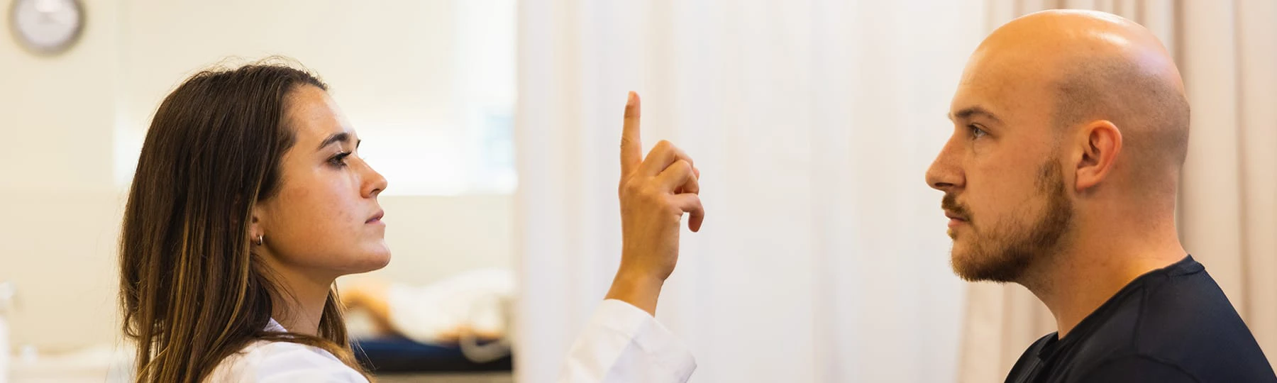 A pre-optometry student holds one hand with her pointer finger up in front of a male patient sitting on a bed.