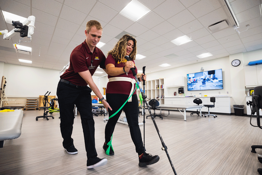 A pre-physical therapy student works with a patient to build strength through walking with a belt and walking sticks.