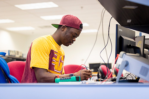 A student in a maroon and gold t-shirt and a baseball hat working on the electronics from inside a computer.