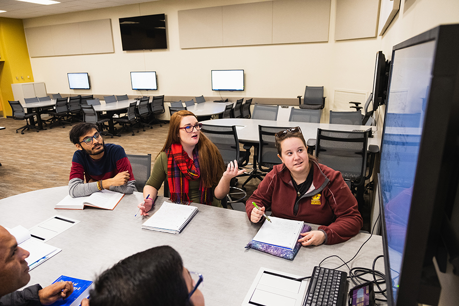 Students sit around a table looking at a monitor on the wall near them.