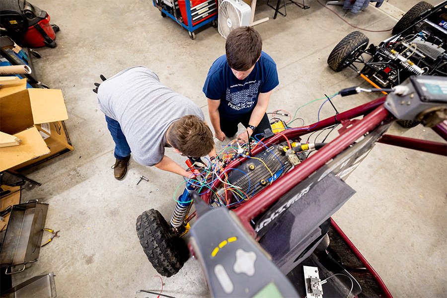 Two students work on the wiring of a off-road vehicle in a open workshop.