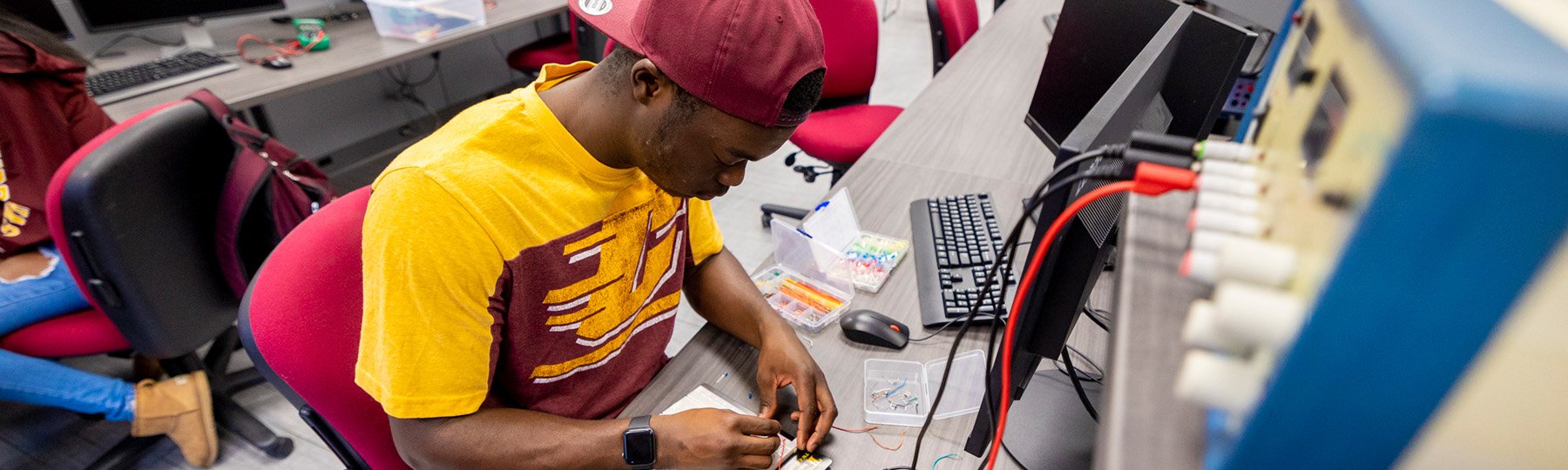 Electrical engineering major in a CMU shirt and hat working on a circuit board in front of electrical equipment.