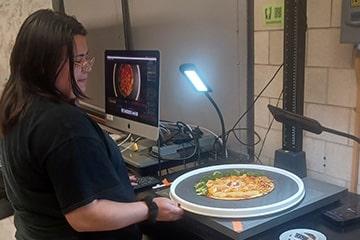 A student holding a pizza on a turntable under a light next to a computer.