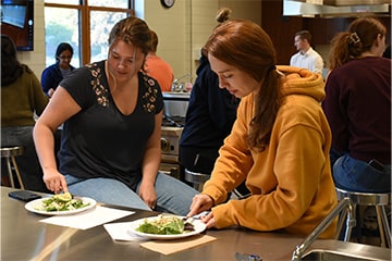 Two students sitting at a stainless steel cooking station with plates of food.