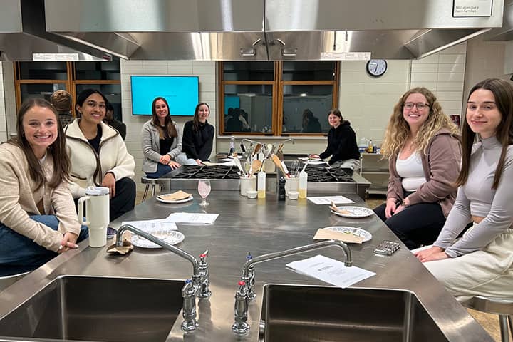 Seven students sitting around a stainless-steel cooking station.