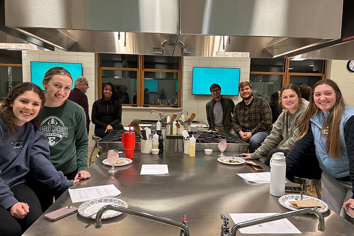Seven students sitting around a stainless-steel cooking station.