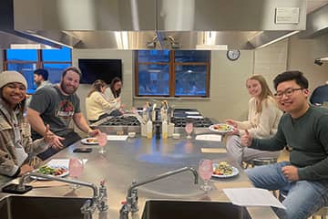 Six students sitting around a stainless-steel cooking station.