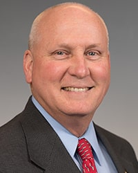 Dr. Greg Zimmerman wearing a black jacket, light blue shirt, and red tie standing in front gray backdrop and looking at the camera.