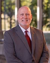 Dr. Greg Zimmerman wearing a black jacket, light blue shirt, and red tie standing in front gray backdrop and looking at the camera.
