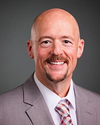 Blaine wears a gray suit jacket and maroon tie while looking at the camera.