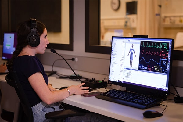 A student wearing headphones sits in the simulation control center at a desk with a computer.