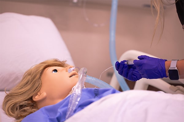 A person in purple gloves holds a syringe near a health simulation mannequin.