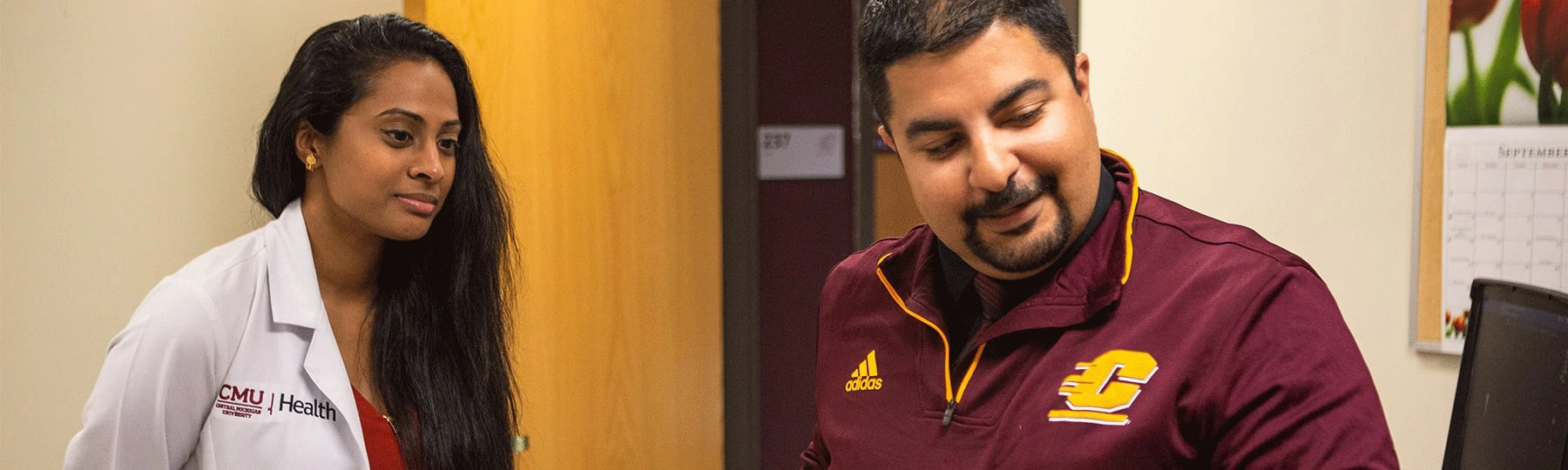 An individual in a white CMU Health lab coat stands next to another individual who is wearing a maroon CMU quarter zip, as they look over a patient in a doctors office.