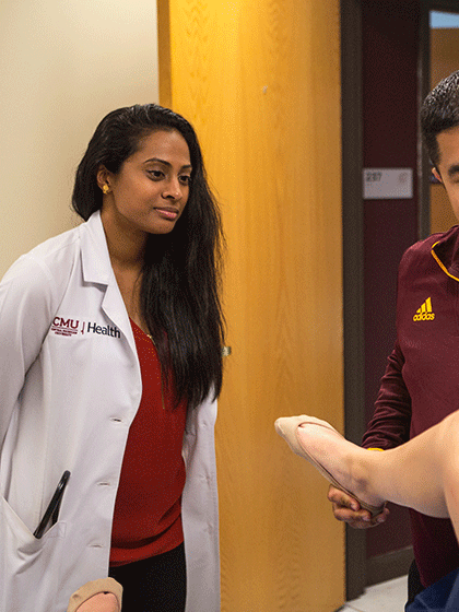 An individual in a white CMU Health lab coat stands next to another individual who is wearing a maroon CMU quarter zip, as they look over a patient in a doctors office.