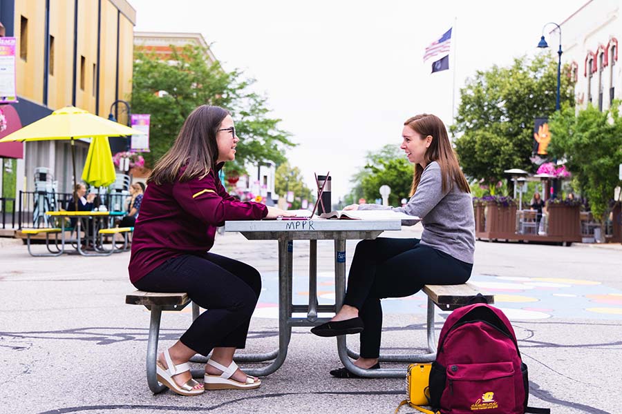 Two female online students sit at a picnic table working on their assignments.