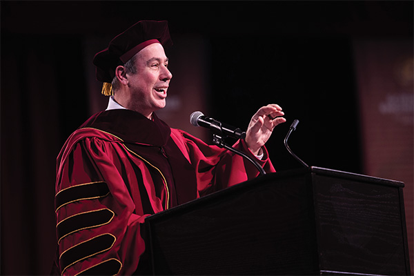 Dr. Neil MacKinnon wearing academic regalia addresses an audience from behind a podium.