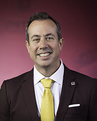 President-Elect Neil MacKinnon wears a maroon suit with a gold tie while posing for a professional head shot against a maroon background.