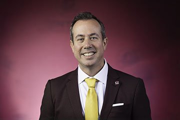 President-Elect Neil MacKinnon wears a maroon suit with a gold tie while posing for a professional head shot against a maroon background.