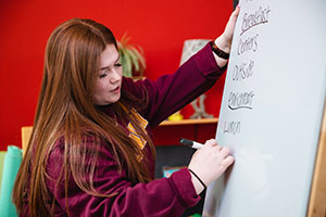 A young woman with long red hair writes out a lesson plan for pre-school children on a large white board.