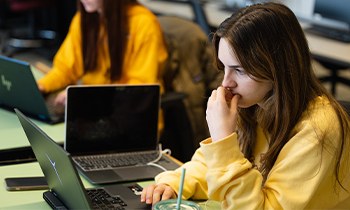 A student in a yellow shirt on her laptop with another student in the background wearing a yellow shirt and working on her laptop as well.