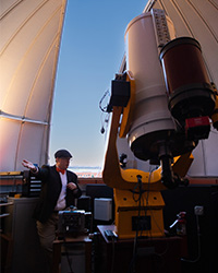 A man in a news cap and bow tie gestures near a telescope looking out into the partially open observatory doors of Brooks Astronomical Observatory.