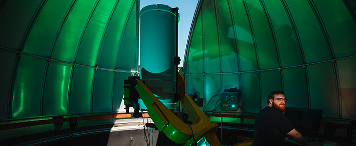 A man wearing glasses stands in Brooks Astronomical Observatory which is partially open with the telescope pointing up.