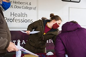 a woman wearing a mask and holding papers helps a women in purple coat at outdoor clinic