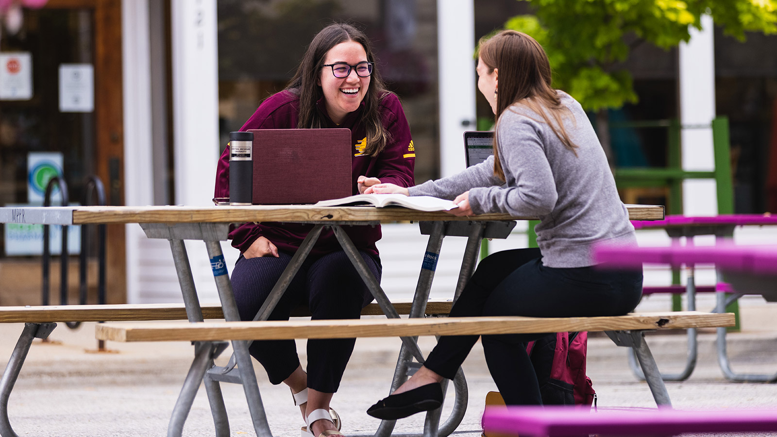 Two women at sitting outside at a restaurant picnic table working together on a project for their online class offered through Central Michigan University.