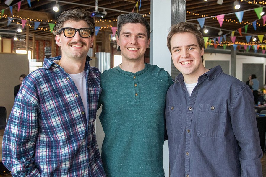 Three young men in casual clothes smile and pose for a picture in a convention center.