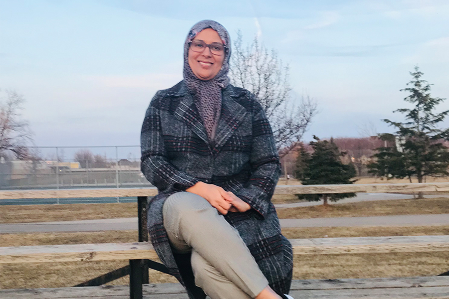 A graduate student wearing a patterned gray hijab sits on a park bench and smiles