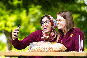 Two girls laughing and smiling as they take a selfie outside. They are both wearing CMU gear, marron sweatshirts and green trees are behind them as they sit at a picnic table with a bag of popcorn on top of the table.