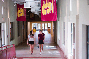 Two girls walk through a residential hall on campus, as they are talking to each other and smiling. There are two flags above them hanging from the ceiling, both are maroon and gold with action C's on them.