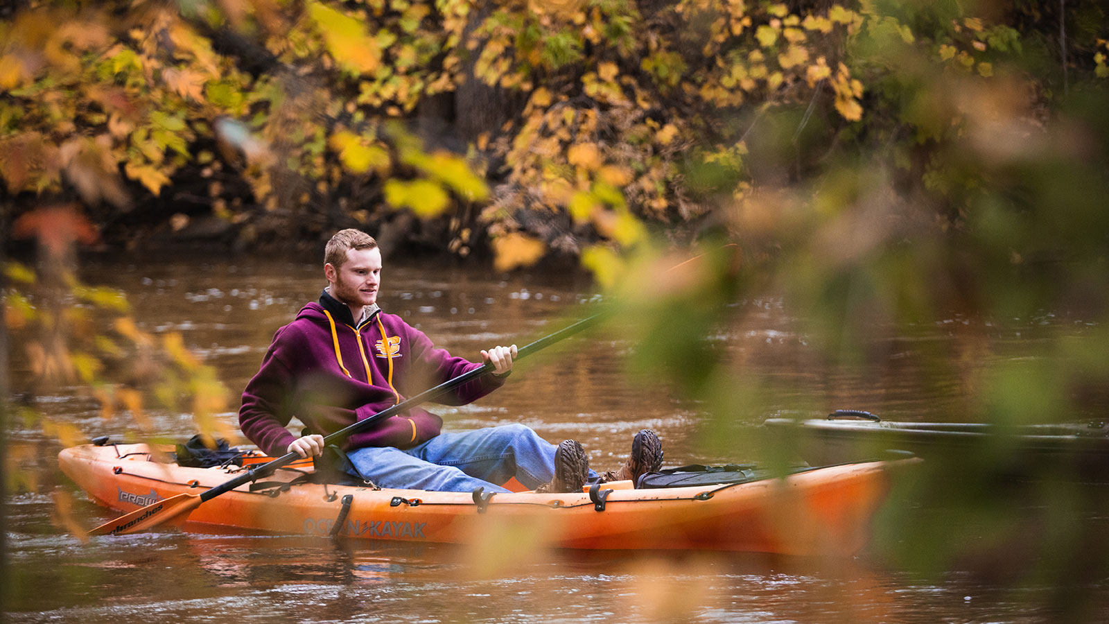 A student kayaks down a river through fall foliage.