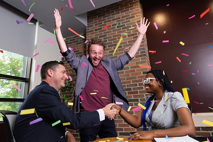 Two people in professional clothes shake hands at a conference table. A person stands in the background cheering in excitement while confetti falls.