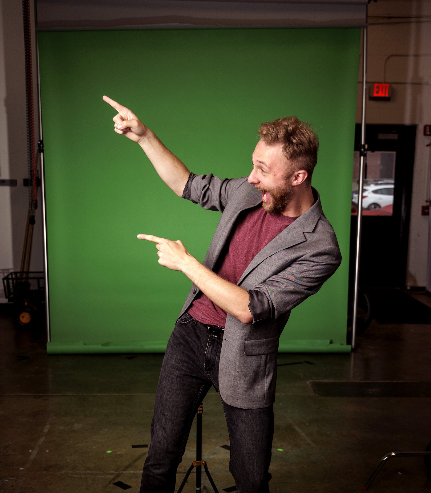 Male student pointing left in front of a studio greenscreen with a dark grey background and green backdrop.