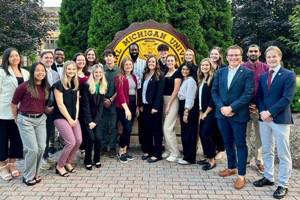 Students in business casual stand together in front of the campus seal.