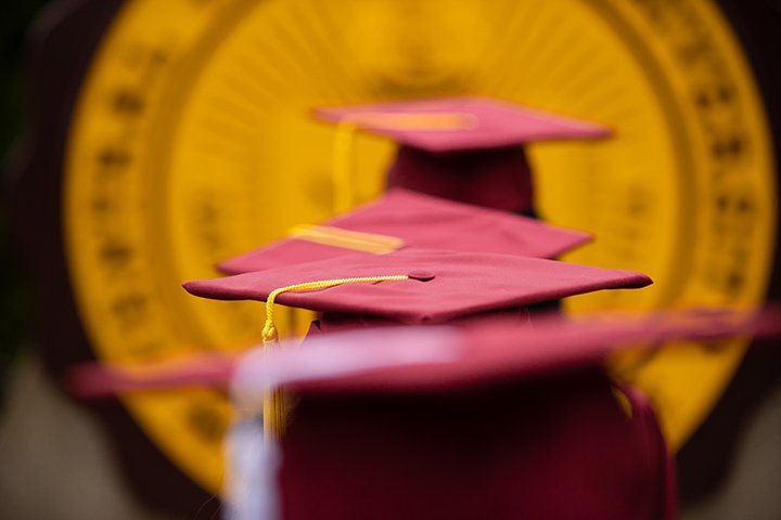 Four CMU students in graduation outfits toss their caps in the air in front of Warriner Hall.