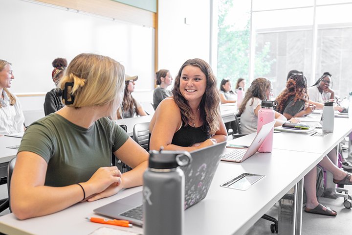 Two students talk to each other while sitting at a classroom desk.