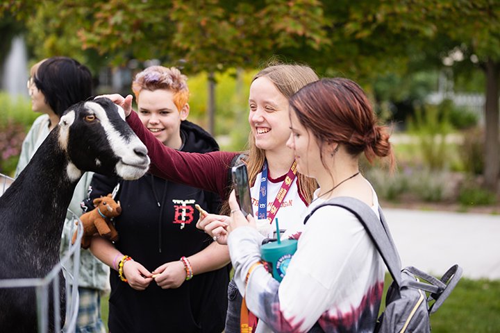 Three CMU students smile and pet a black and white goat.