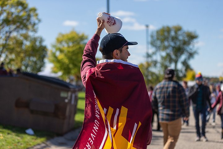 A CMU student draped in a CMU flag has his back to the camera while raising his left arm in the air.