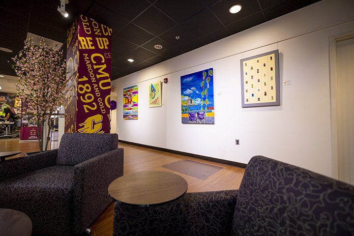 Two dark-colored chairs and brown table in the foreground of a room with artworks on canvas hanging on the wall in the background.