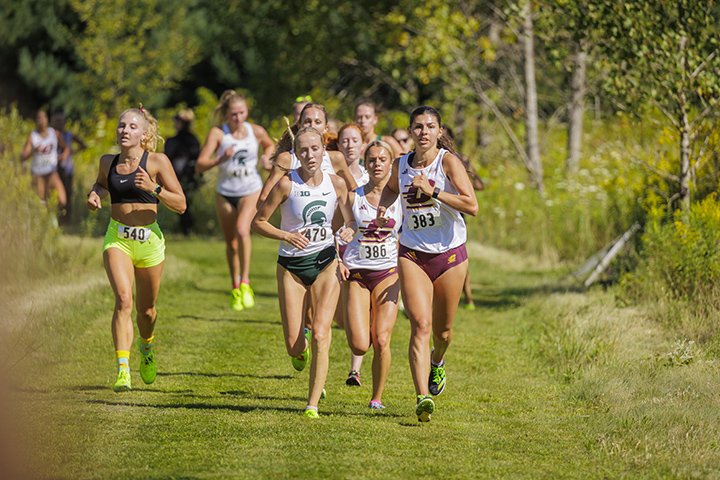 A group of women's cross country runners race through the woods. The front three runners include two wearing CMU uniforms and one wearing a Michigan State University uniform.
