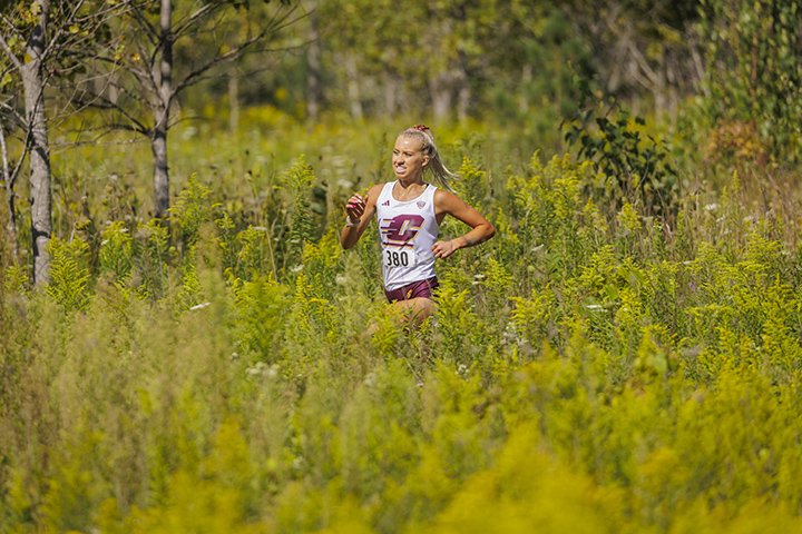 A CMU women's cross country athlete runs through the woods, surrounded by shrubs, trees and tall grass.