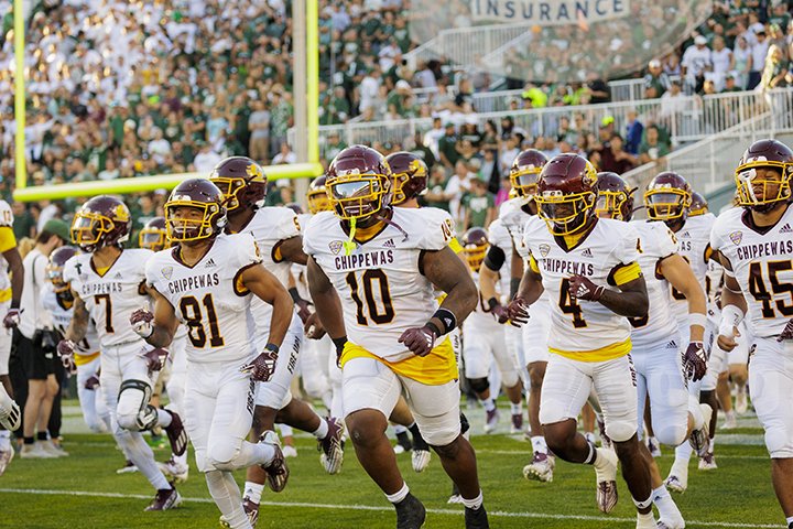 CMU football players run onto the field at Michigan State University wearing white uniforms and maroon helmets as the crowd cheers in the background.